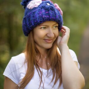 Une femme porte un bonnet en maille colorée avec pompon, dans un décor naturel verdoyant.